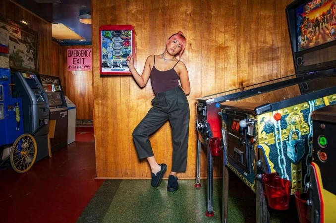 Young woman with pink hair leans against a wood panel wall near pinball and vending machines in a vintage-style game room indoors.