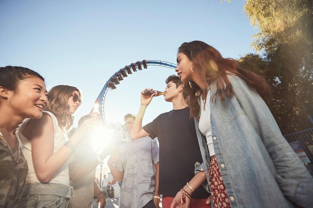 Group of diverse young adults enjoying drinks and camaraderie at an amusement park with a roller coaster in the background on a sunny day.