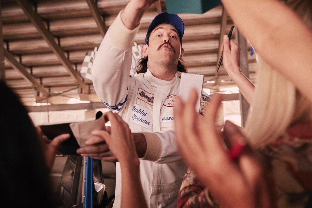 Adult male in racing suit celebrated by group in garage setting, holding papers with blue cap. Racing, team, indoors, celebration.