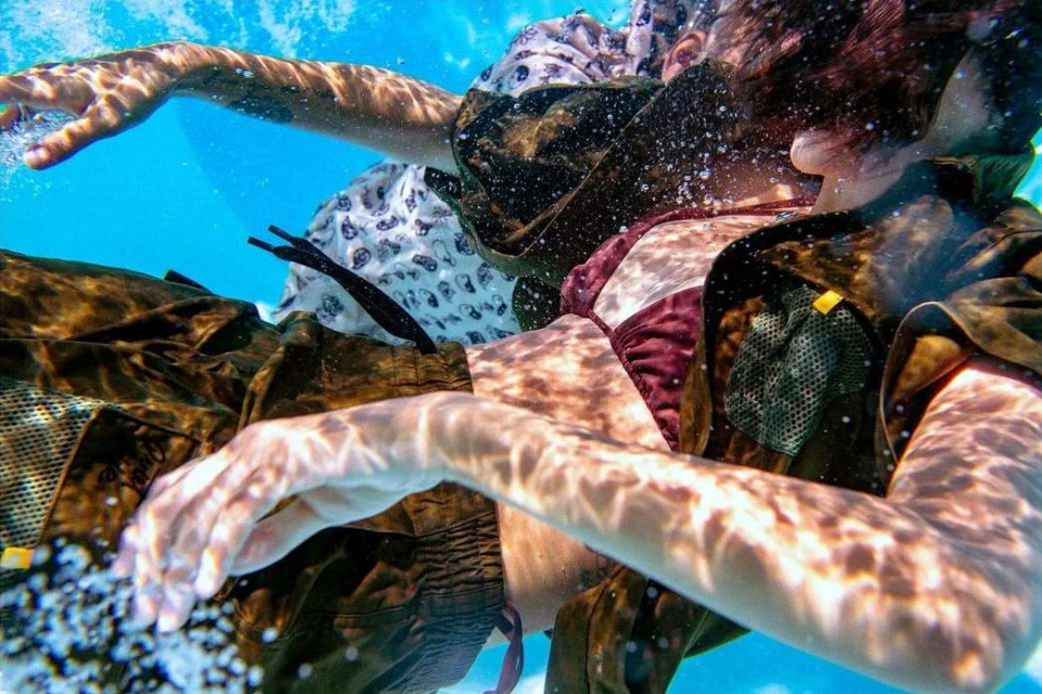 Underwater shot of two people in casual attire, enjoying the clear blue water with sunlit patterns. Shirt and swimwear are visible.