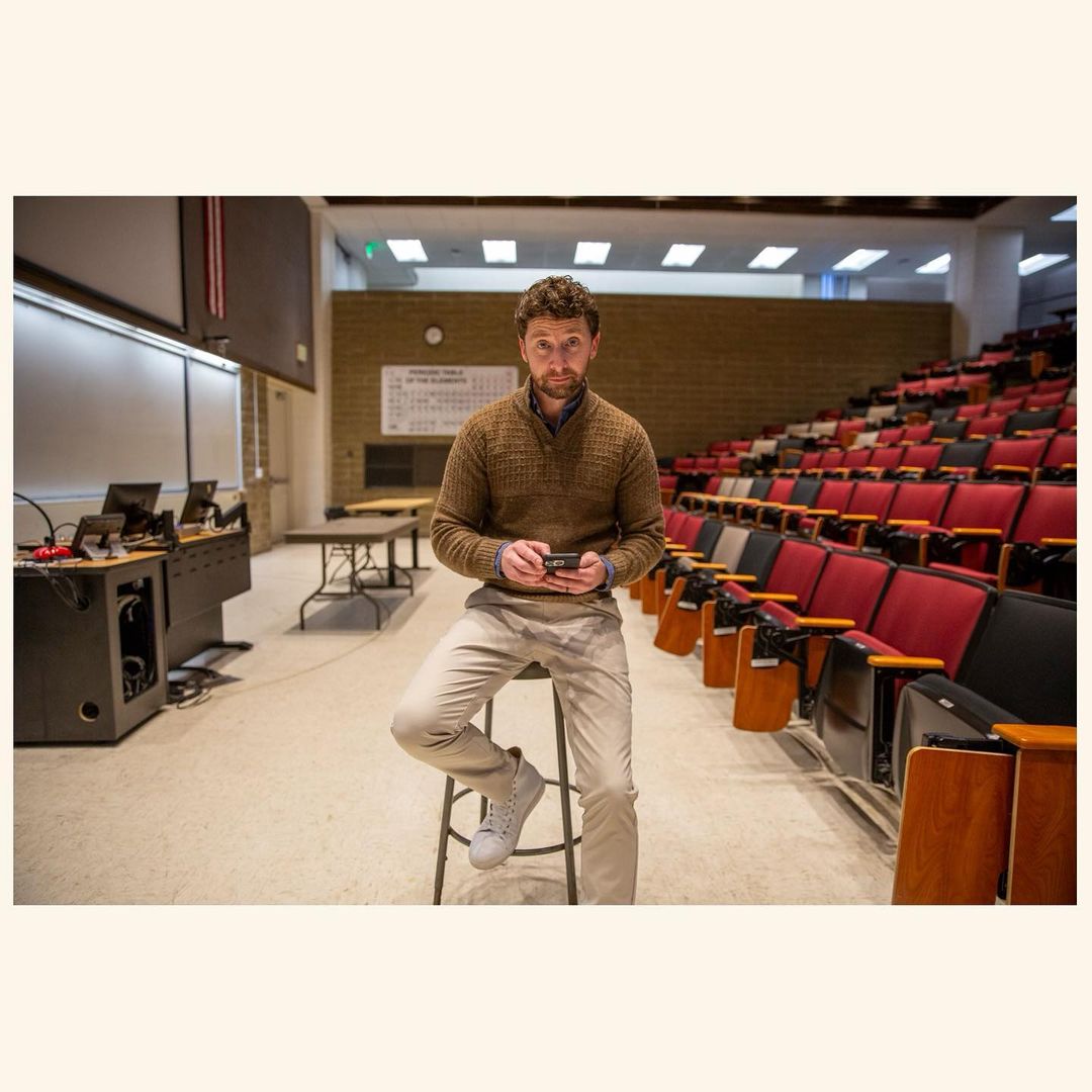 Man sitting on stool in auditorium holding phone, wearing brown sweater, khakis, and sneakers, looking at the camera.