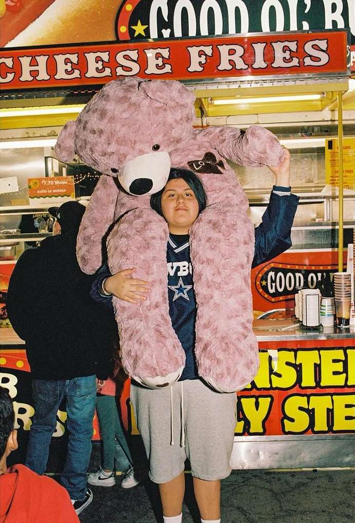Woman with long dark hair carrying large pink teddy bear, wearing a Dallas Cowboys top and grey shorts in front of food stand.