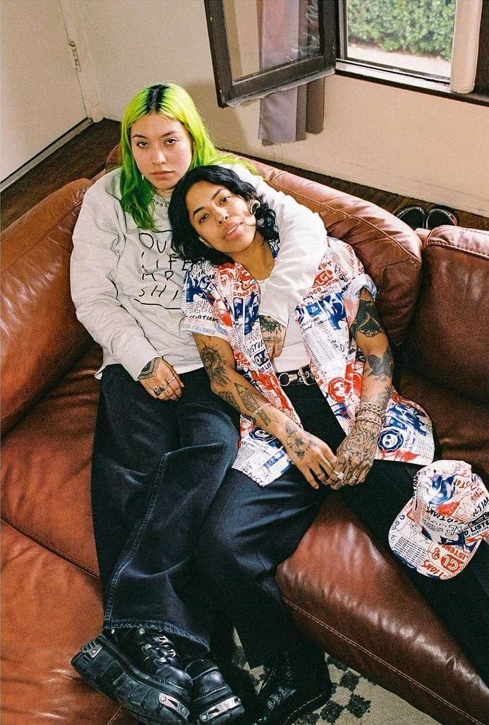 Two women pose intimately on a brown leather couch in a brightly lit indoor setting with modern casual fashion and tattoos.