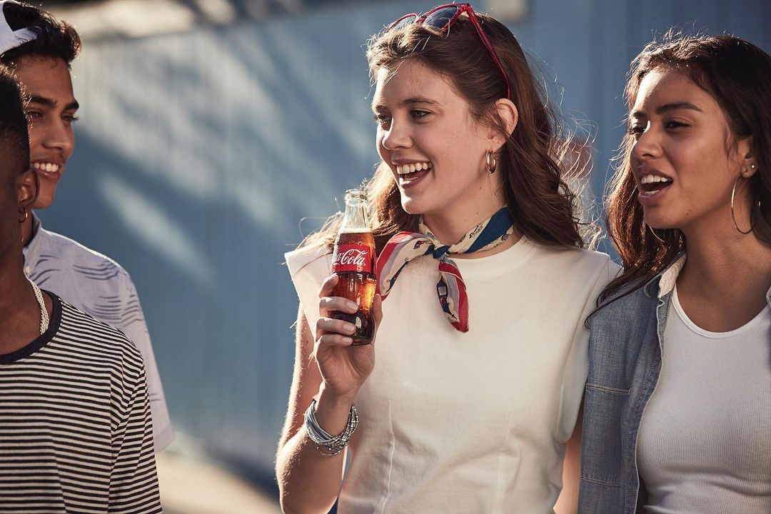 A group of young adults stand outside, smiling, with a woman holding a Coca-Cola bottle, creating a casual, cheerful, and refreshing scene.