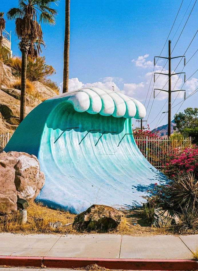 Sculptural wave art installation in a desert setting with palm trees against a clear blue sky, creating a whimsical and surreal scene.