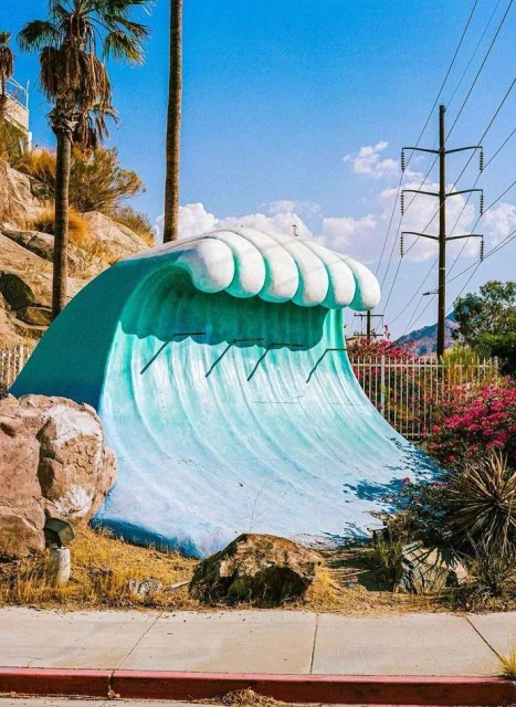 Sculptural wave art installation in a desert setting with palm trees against a clear blue sky, creating a whimsical and surreal scene.