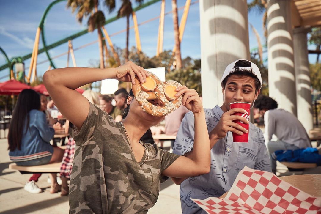 Two people at an outdoor cafe: one woman holds a large pretzel with cheese in front of her face, while a man drinks from a Coca-Cola cup.