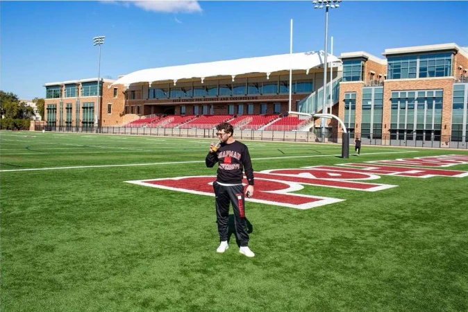 Adult male stands on a green football field in front of a stadium under a blue sky on a sunny day, holding a beverage.