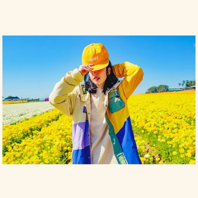 Woman in a yellow ball cap and colorful jacket standing in a field of yellow flowers under a blue sky on a sunny day. 