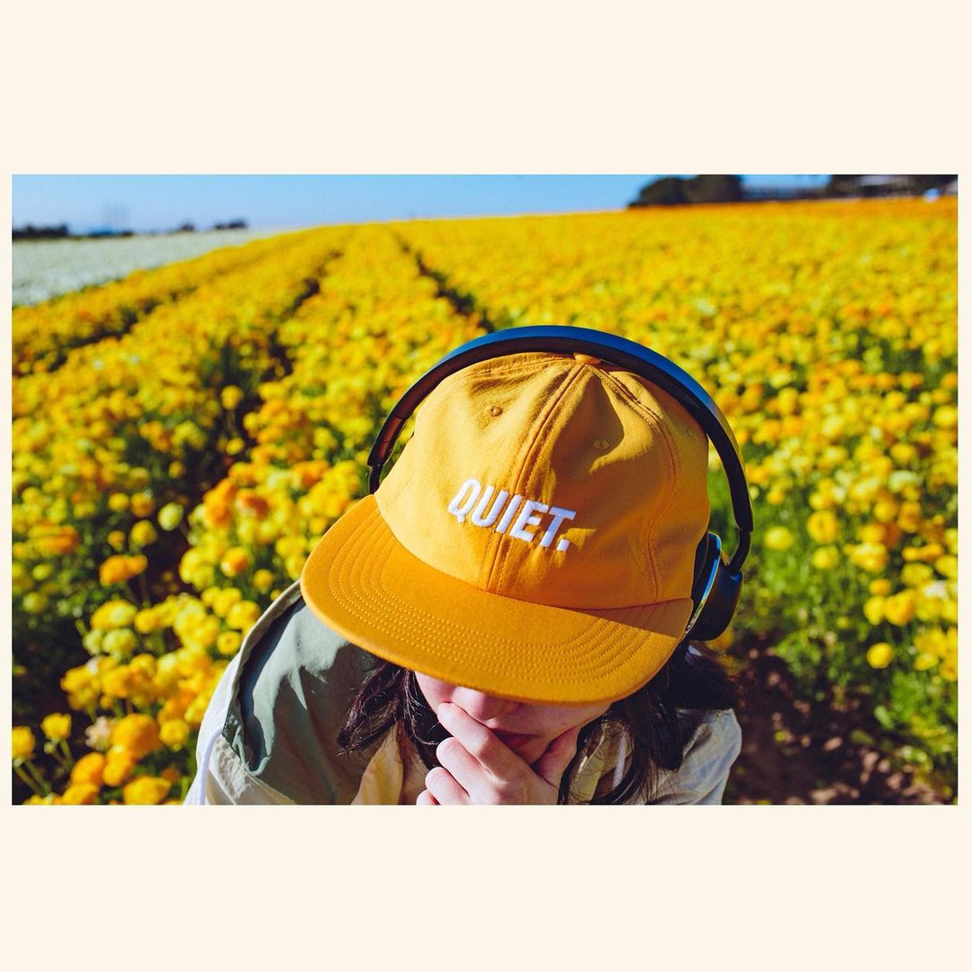 A woman wearing a yellow baseball cap and headphones stands in a vibrant field of yellow flowers under a sunny blue sky.