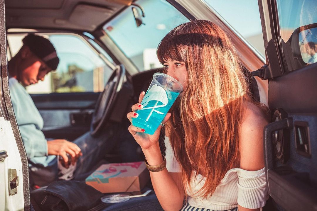 Woman drinks blue beverage in car with another person visible in background; interior shot with natural lighting, and a casual mood.