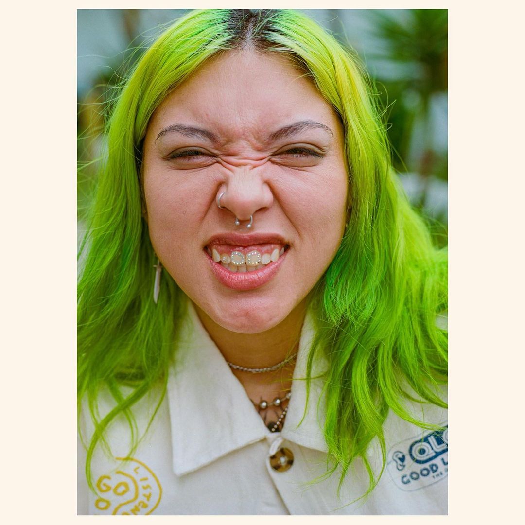 Portrait of a woman with green hair, a nose ring, and a white jacket, set against a blurred outdoor background with green foliage.