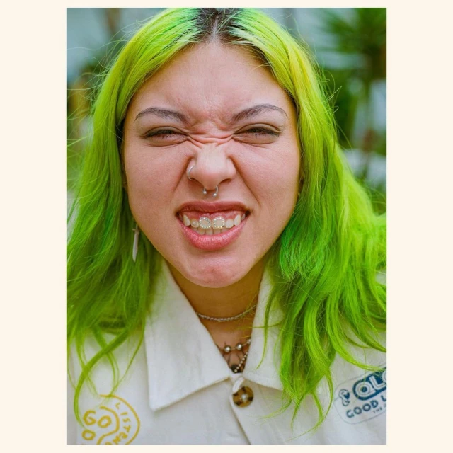 Portrait of a woman with green hair, a nose ring, and a white jacket, set against a blurred outdoor background with green foliage.