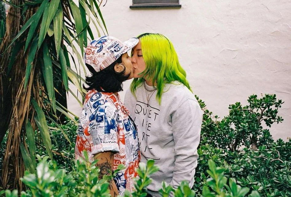 Two women embrace and kiss outdoors, one with bright green hair and the other wearing a patterned shirt and hat, surrounded by greenery.