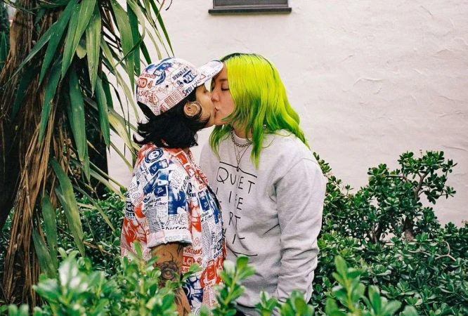 Two women embrace and kiss outdoors, one with bright green hair and the other wearing a patterned shirt and hat, surrounded by greenery.
