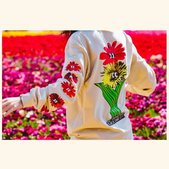 Person in flower field wearing a light beige sweatshirt with cartoon flower graphics on the back and sleeves, arms extended.