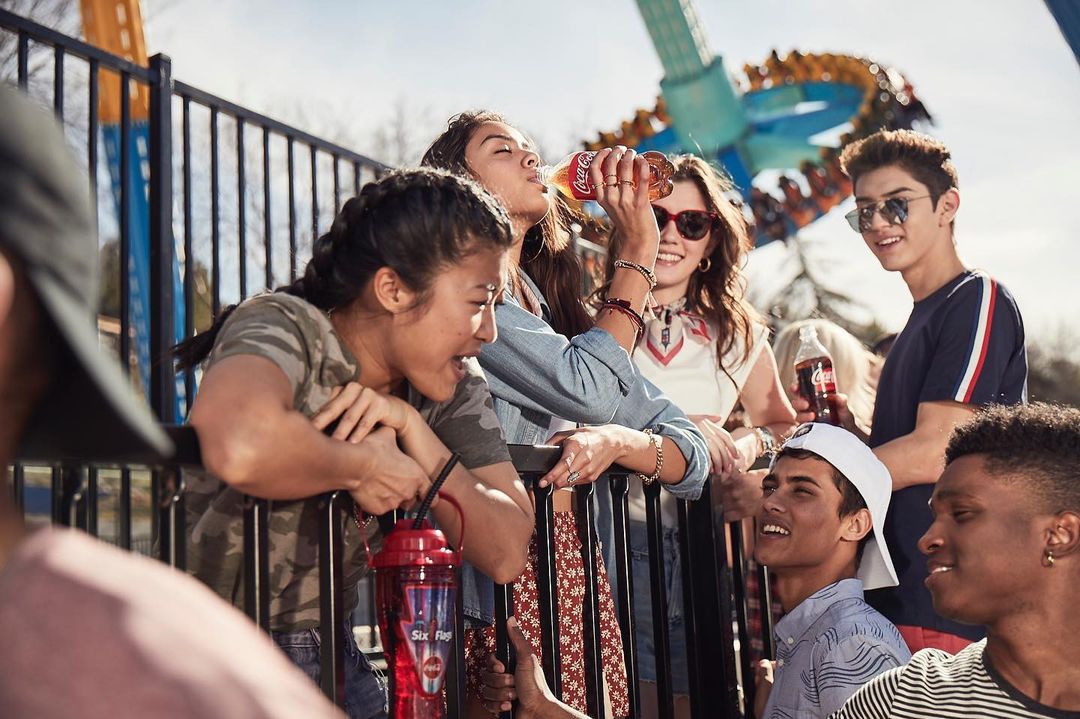Group of young adults drinking Coca-Cola, standing behind a metal fence in an amusement park with a high-thrill ride in the background.