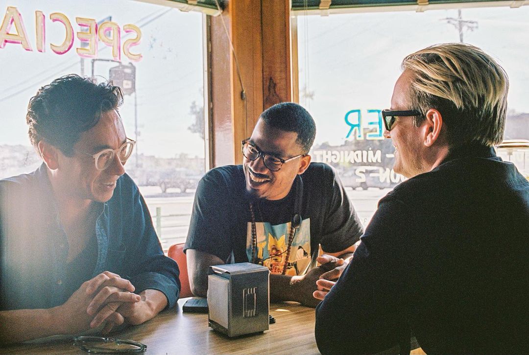 Three men are sitting at a table inside a diner, engaged in conversation. The men are smiling and the lighting is warm and inviting.