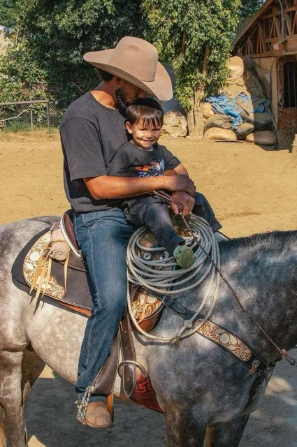 Father and child wearing cowboy hats riding a gray horse outdoors with trees and a shed in the background on a sunny day.