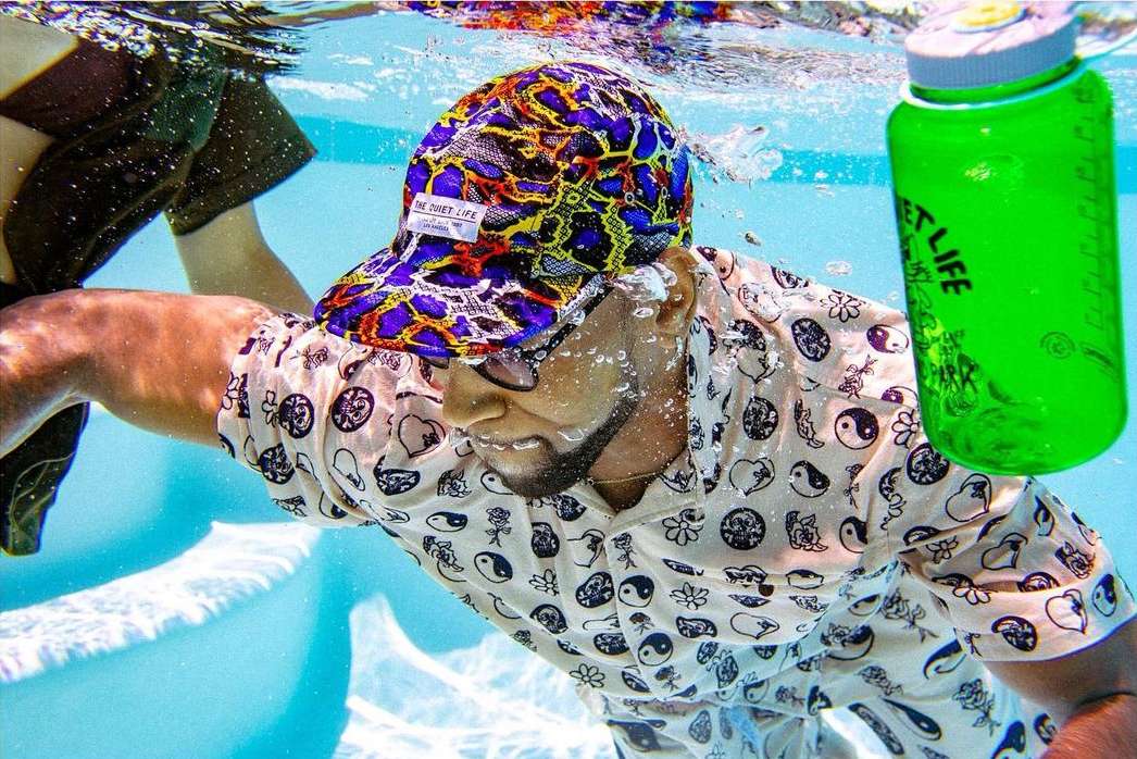 Man with patterned cap and shirt swims underwater in a pool, holding a green water bottle. Swimming and water sports.