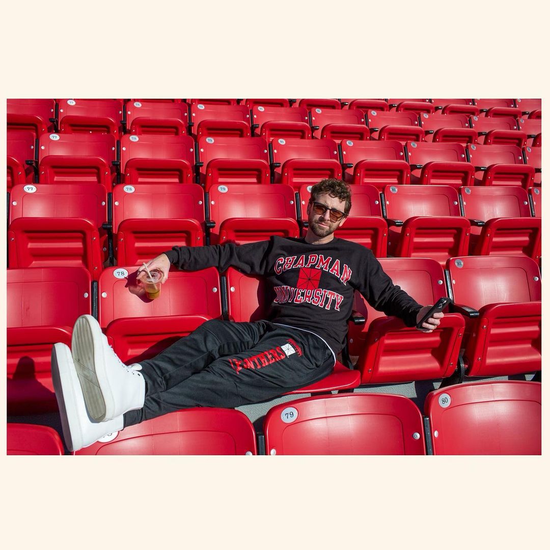 Man sits in a stadium wearing a Chapman University sweatshirt and sweatpants, sunglasses, and white shoes, holding a drink.