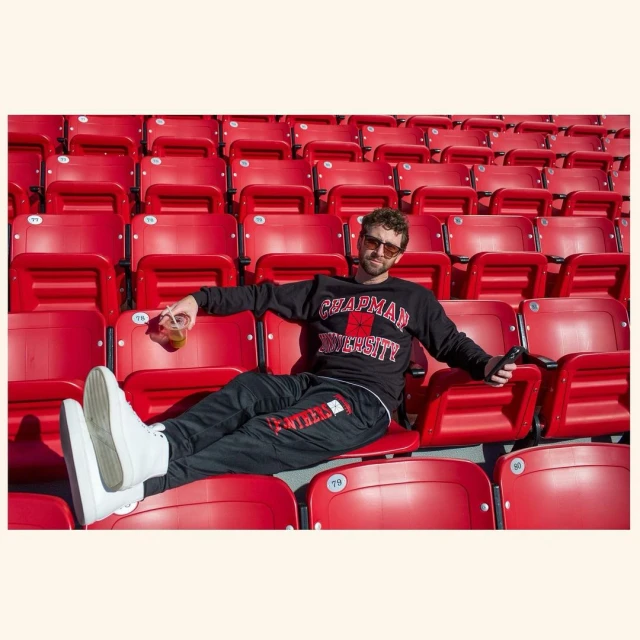 Man sits in a stadium wearing a Chapman University sweatshirt and sweatpants, sunglasses, and white shoes, holding a drink.