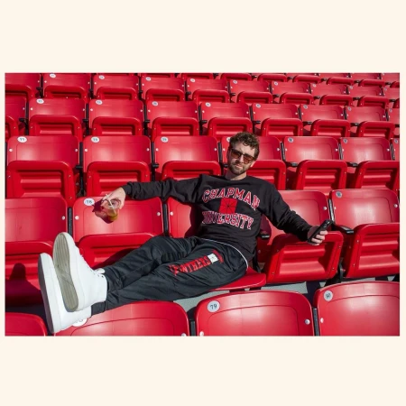 Man sits in a stadium wearing a Chapman University sweatshirt and sweatpants, sunglasses, and white shoes, holding a drink.