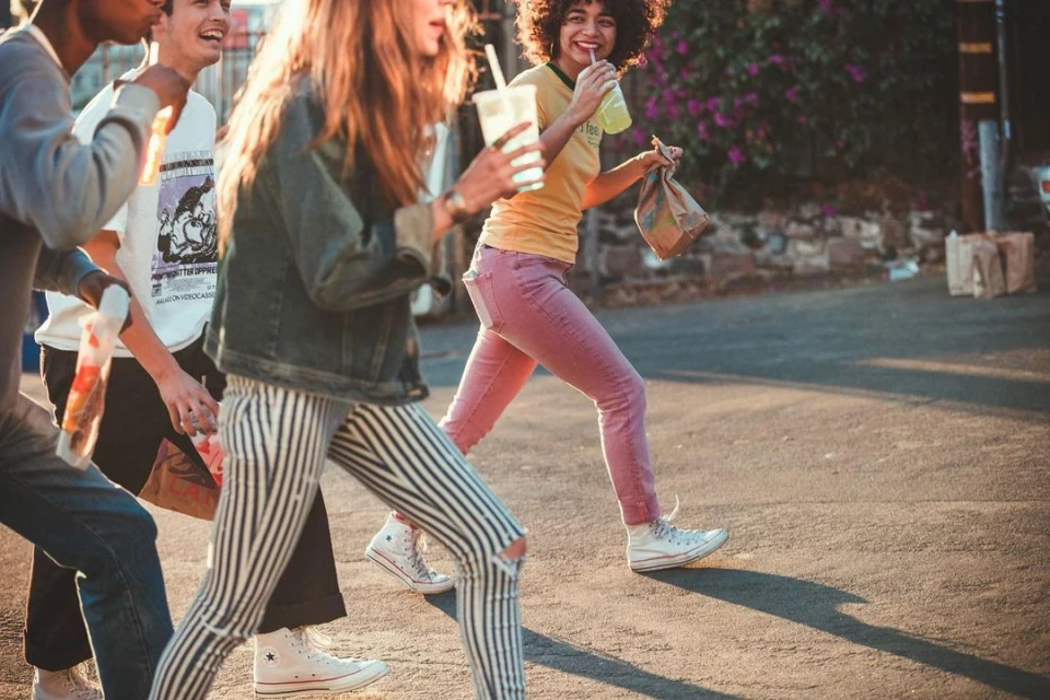 A diverse group of young adults strolls down a sunlit street, holding drinks and food bags, clad in fashionable casual attire with vintage vibes.