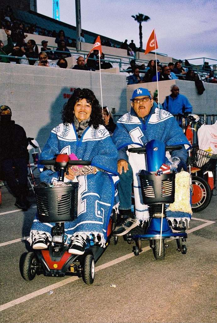 Elderly couple in matching blue outfits and Los Angeles Dodgers hats ride mobility scooters at an outdoor event with stadium seating.
