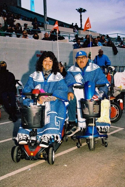 Elderly couple in matching blue outfits and Los Angeles Dodgers hats ride mobility scooters at an outdoor event with stadium seating.