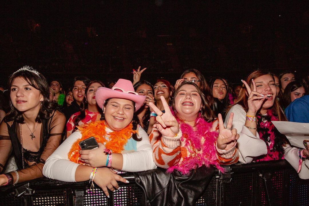 A crowd of young adults are standing behind a metal barrier at an indoor concert, smiling and making peace signs with their fingers.