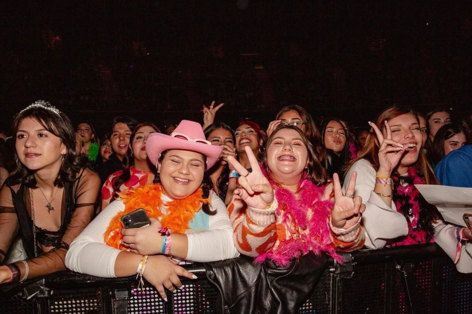 A crowd of young adults are standing behind a metal barrier at an indoor concert, smiling and making peace signs with their fingers.
