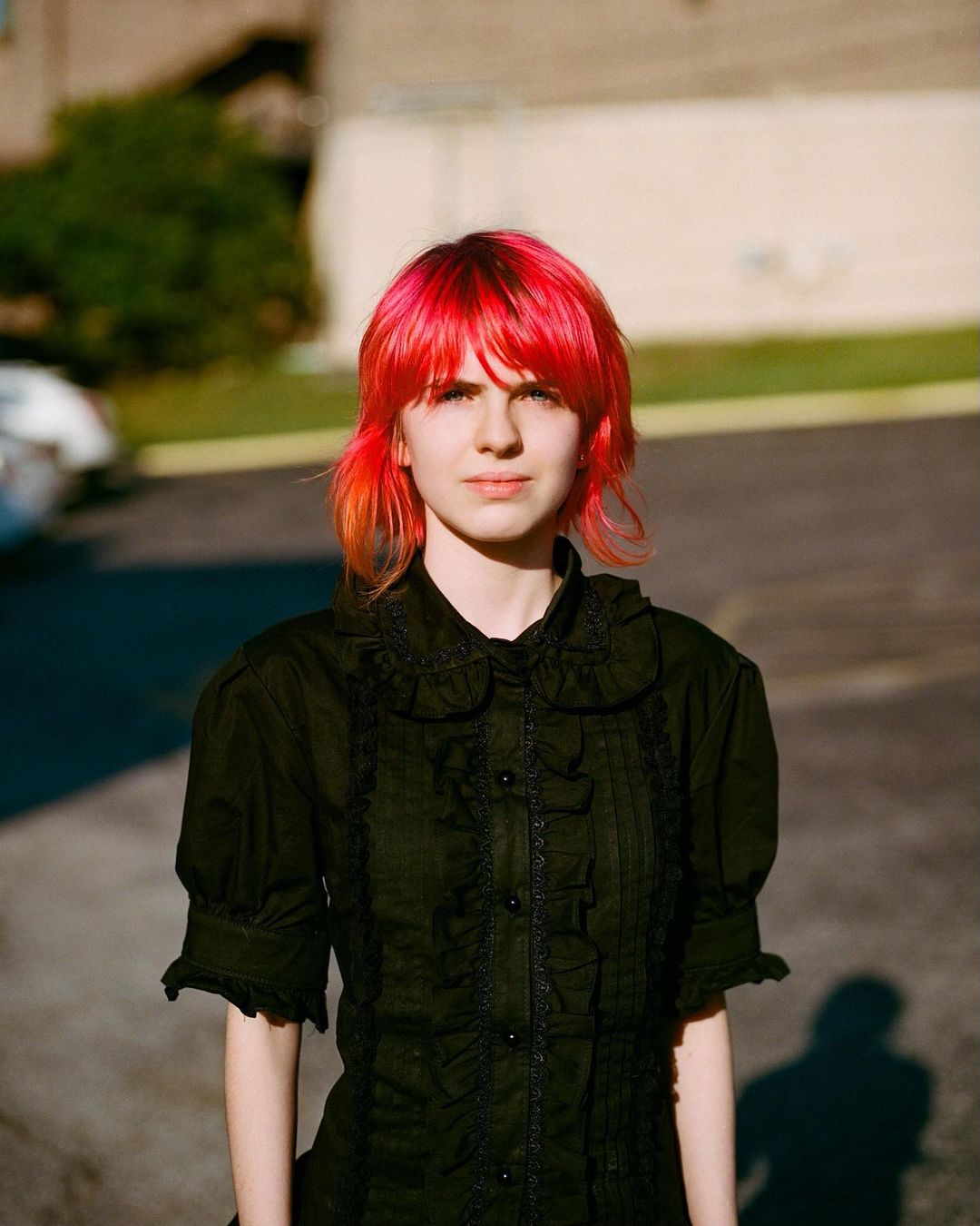 Young woman with short red hair and black ruffled shirt stands in urban daylight, creating a modern and edgy fashion portrait.