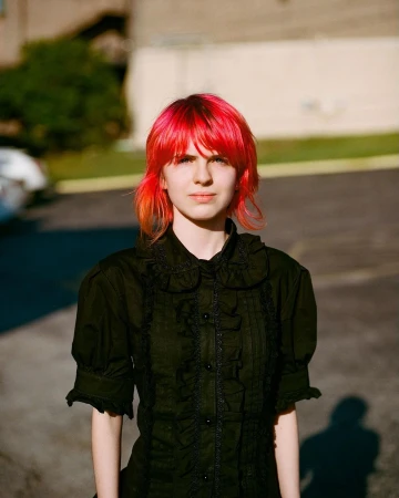 Young woman with short red hair and black ruffled shirt stands in urban daylight, creating a modern and edgy fashion portrait.