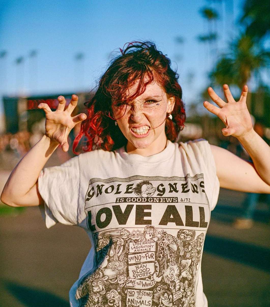 A young woman with red hair and freckles poses outdoors, wearing a graphic tee and making expressive hand gestures in sunlight.