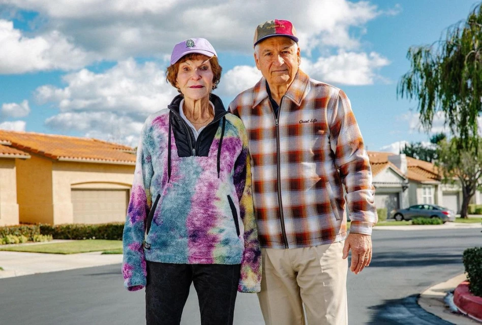An elderly couple stands on a suburban street, posing in front of houses on a sunny day with a blue sky and scattered clouds.