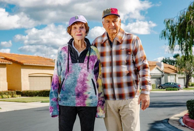 An elderly couple stands on a suburban street, posing in front of houses on a sunny day with a blue sky and scattered clouds.
