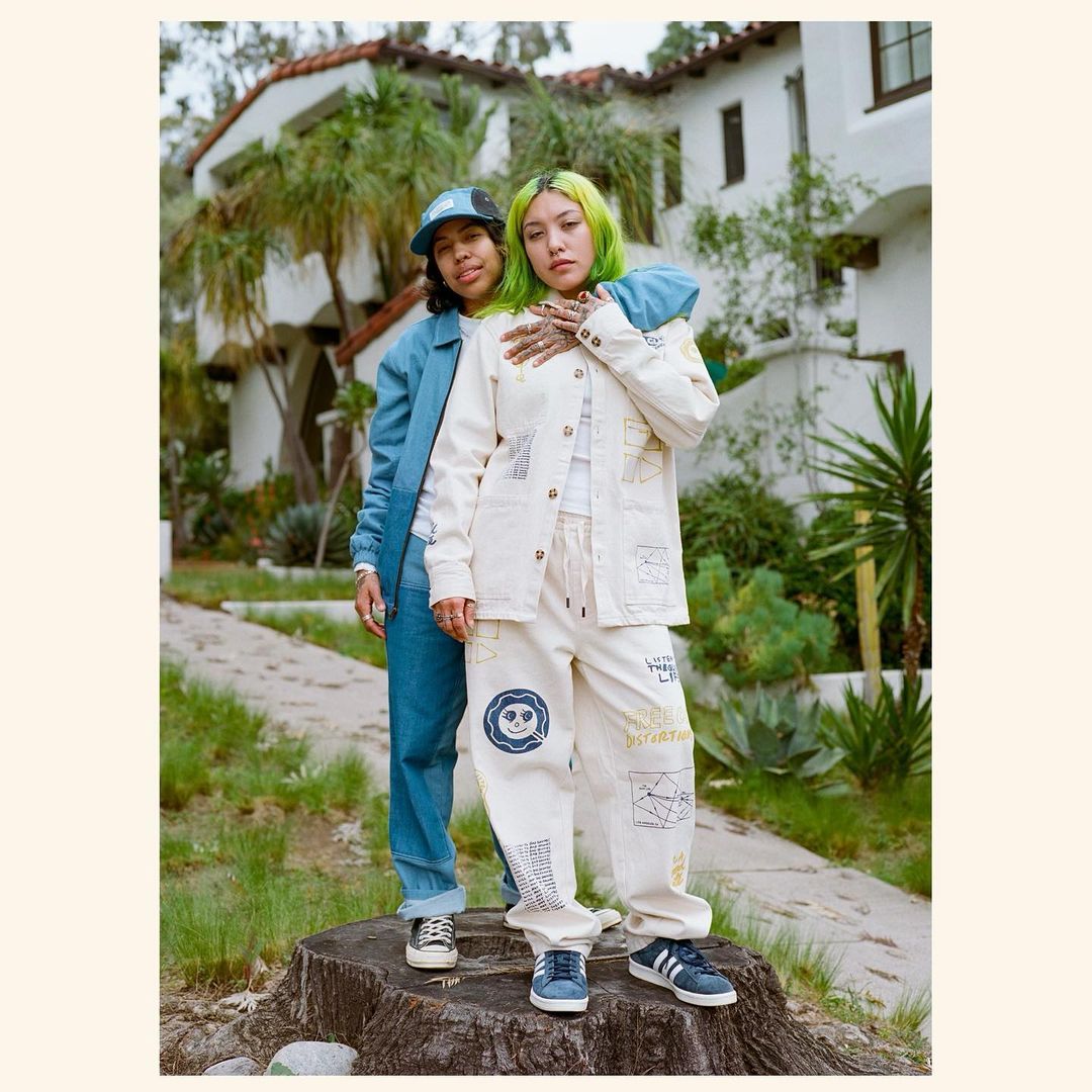 Two women pose outdoors near a building in denim and cream-colored clothing with graphic patterns. One has bright green hair.