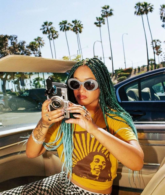 A young adult woman takes a photo in a car using a digital camera, wearing sunglasses and a yellow t-shirt with braided hair.
