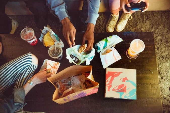 Overhead shot of four individuals gathered around a dark wooden table with food items from Taco Bell and a gaming controller.