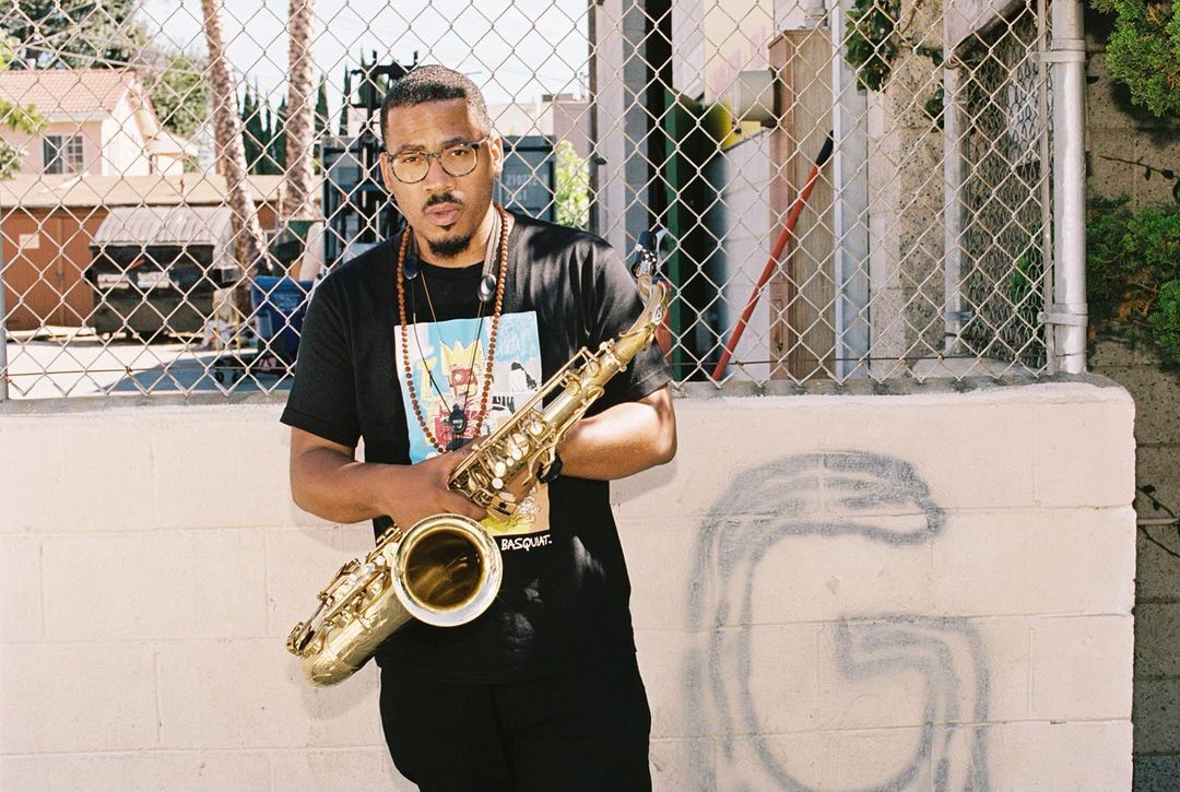 African-American man stands outdoors holding a saxophone, wearing a graphic tee and beaded necklaces, medium shot, urban setting.
