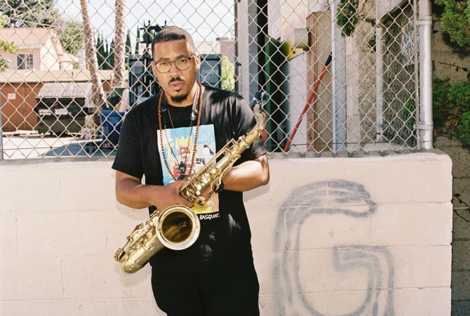 African-American man stands outdoors holding a saxophone, wearing a graphic tee and beaded necklaces, medium shot, urban setting.