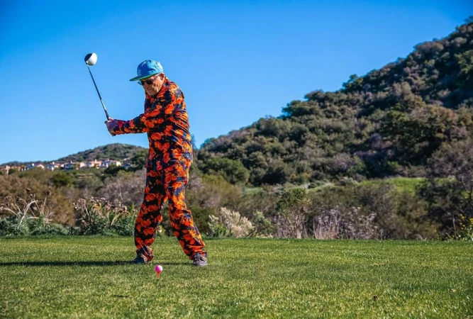 Elderly man in patterned jumpsuit swinging golf club on green golf course on a sunny day, mountain and blue sky background.