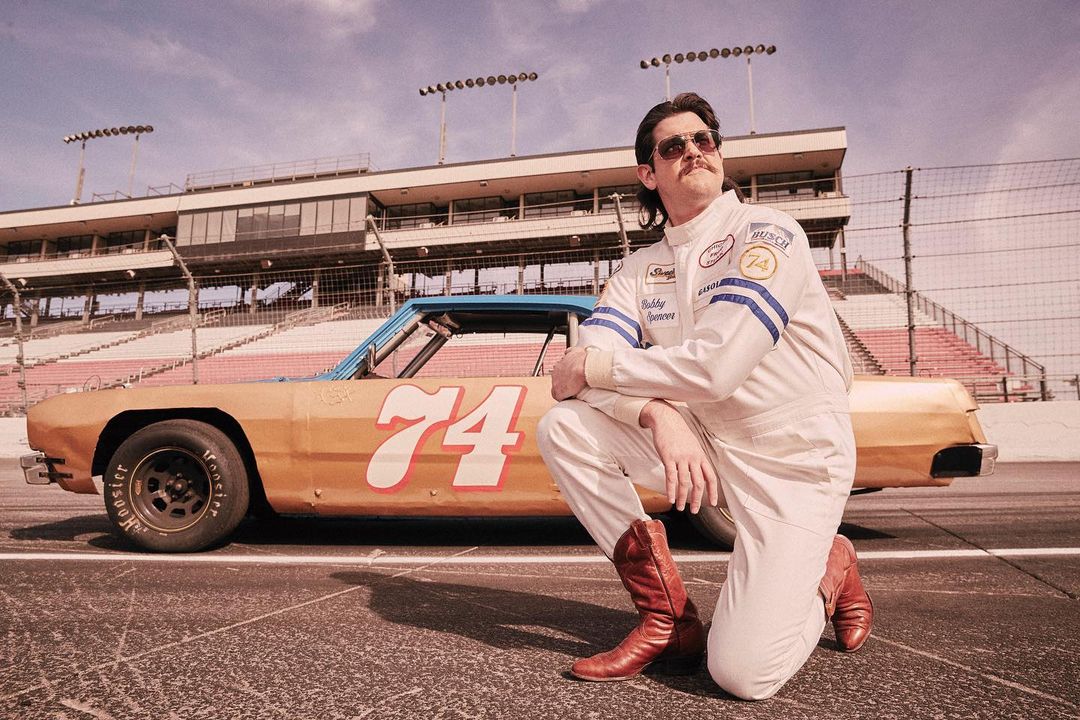Man in racing suit kneels by vintage race car on track, grandstands behind. Daytime outdoor shot with golden-era color palette.