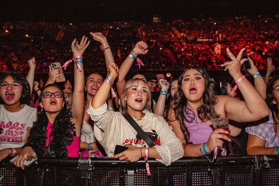 Excited crowd of young adults raises hands at a music event, with bright stage lighting and a sea of faces visible in the background.
