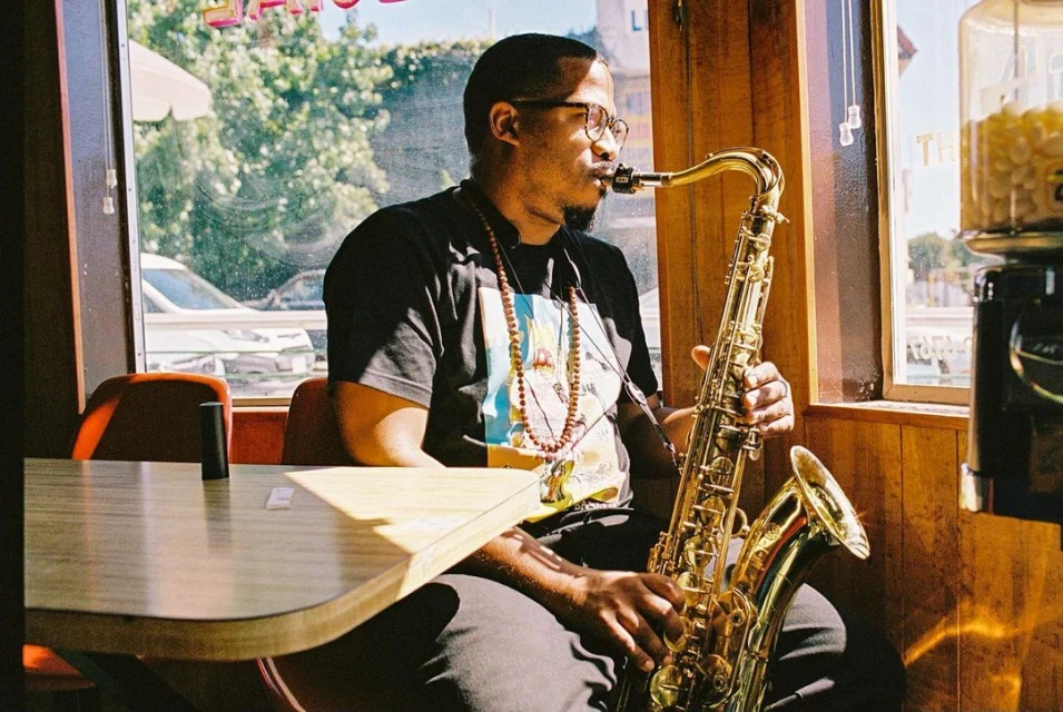 Man with glasses plays saxophone at table in wood-paneled cafe near window on sunny day, cars visible on street outside.