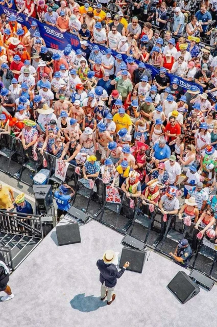 Large crowd wearing blue hats gathers around a stage with a man in a suit. There are signs and sponsor logos in the background.