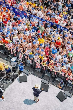 Large crowd wearing blue hats gathers around a stage with a man in a suit. There are signs and sponsor logos in the background.