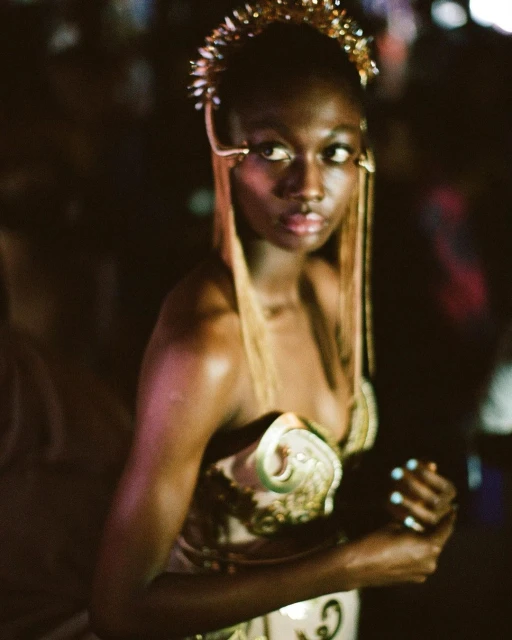 Stunning dark-skinned woman with ornate gold headpiece and bustier gazes confidently in a dimly lit environment, showcasing fashion and beauty.