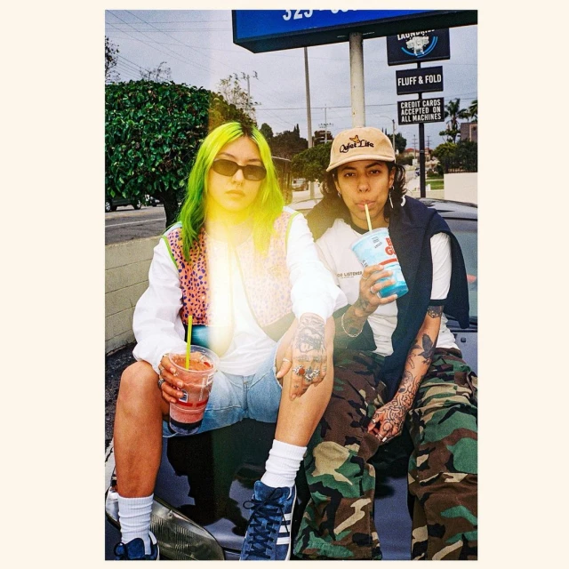 Two young adults sit on a car with drinks outside a laundromat under overcast daylight, wearing casual street fashion with tattoos.